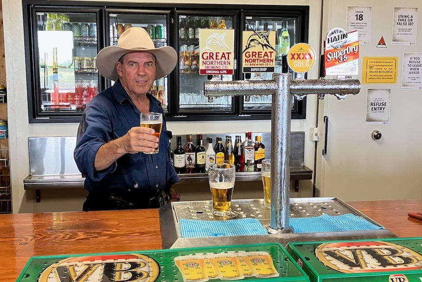A man in a work shirt and large hat stands behind a bar, holding up a fresh cold beer.