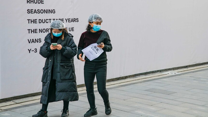 Two women in long puffy jackets with facemasks and hairnets walk on a street holding their smartphones.
