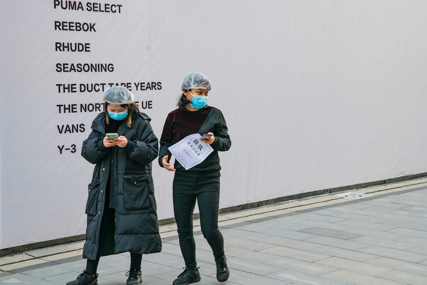 Two women in long puffy jackets with facemasks and hairnets walk on a street holding their smartphones.