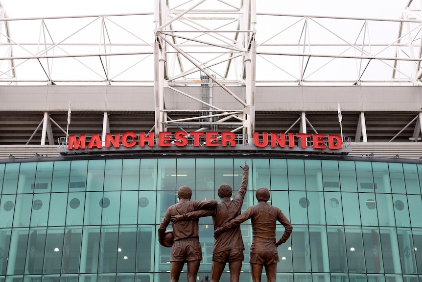 Una statua di tre ragazzi che si abbracciano da dietro e guardano lo striscione del Manchester United su un edificio