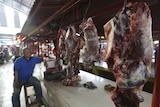 A butcher leans on a stool by a stall lined with meat hanging on hooks at covered markets.