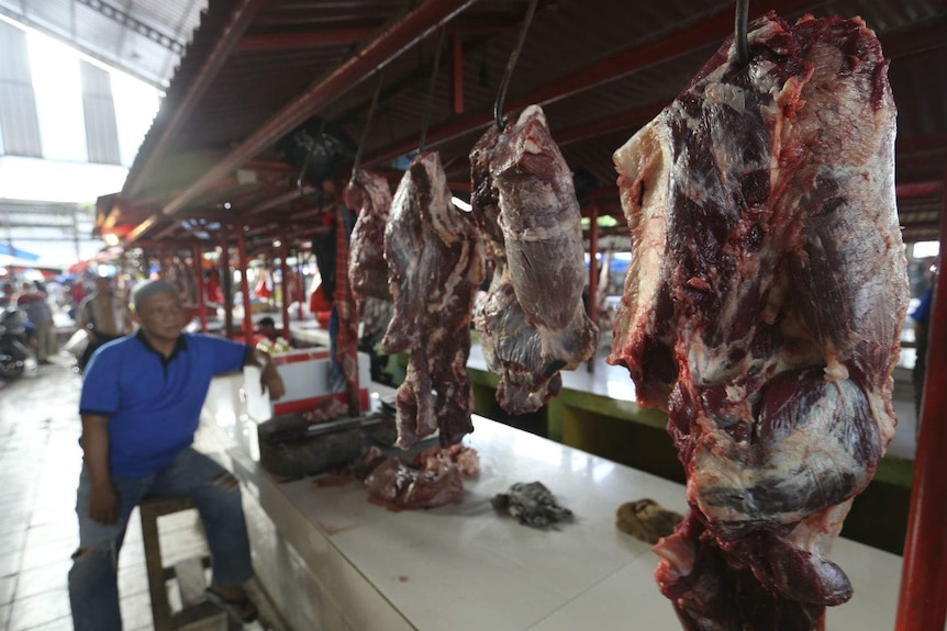A butcher leans on a stool by a stall lined with meat hanging on hooks at covered markets.