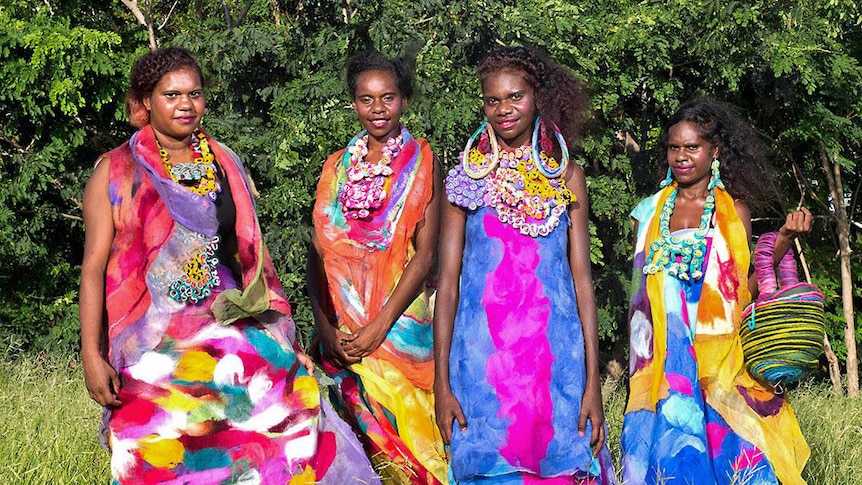Four Indigenous women stand in a field wearing bright coloured felt and silk garments