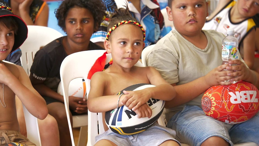 A young, smiling Indigenous boy sits in a chair holding a rugby league ball, surrounded by other sitting Indigenous children