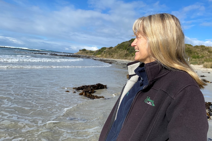 A woman with long blond hair in polar fleece jacket stands on secluded beach in sunny weather smiling looking out to sea