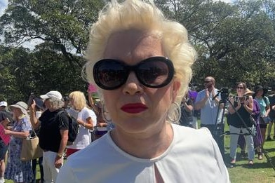 Blonde woman in dark glasses and red lipstick looking into the camera with a park in the background 