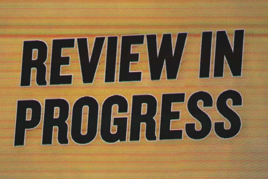 A sign in big black type on a yellow background on an electronic scoreboard shows the words "Review in progress".