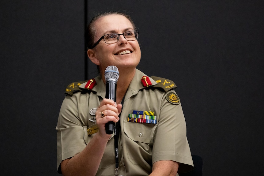 A woman in army uniform with glasses holding a microphone and smiling while she looks up to the right