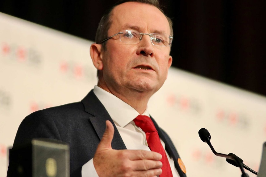 Mark McGowan wearing a suit, red tie and glasses, speaks on stage behind a lectern.