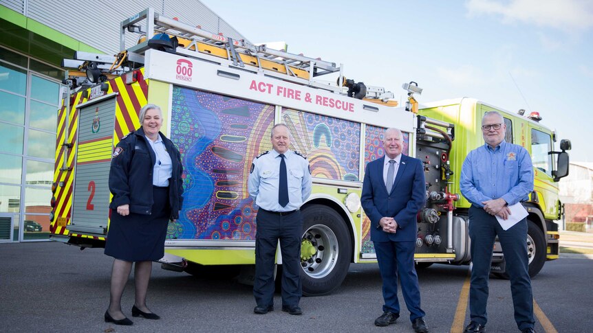 The four people stand smiling at the camera, a fire truck behind them.