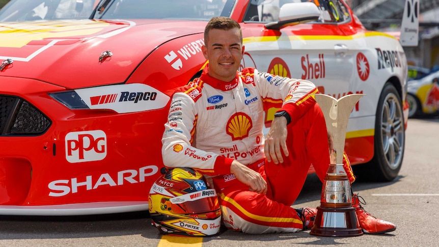 A driver sits on the ground in front of a V8 car with the Supercars championship trophy next to him.