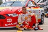 A driver sits on the ground in front of a V8 car with the Supercars championship trophy next to him.