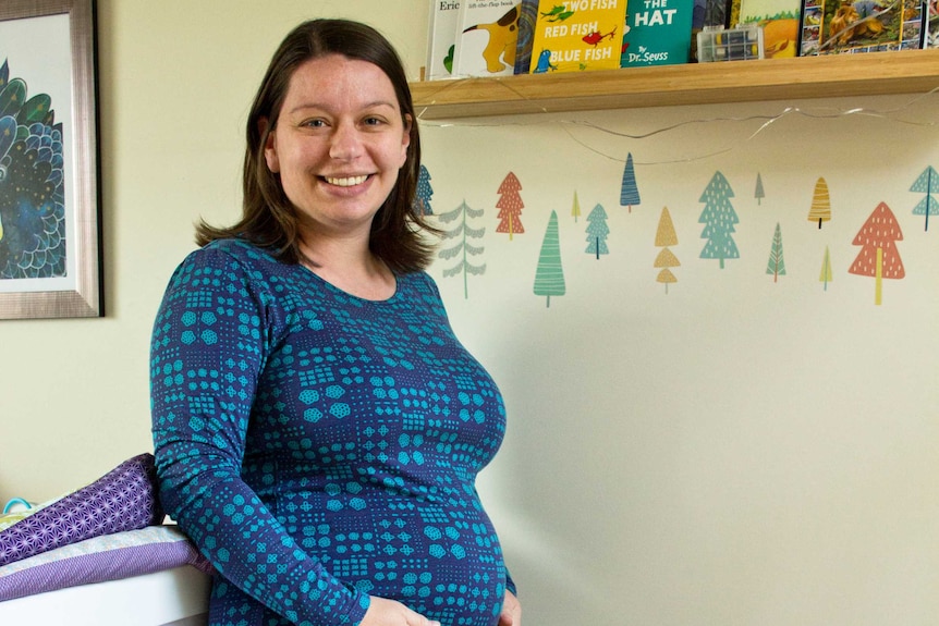 A pregnant woman standing in a nursery holding her belly with kids books on a shelf behind her.