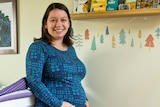 A pregnant woman standing in a nursery holding her belly with kids books on a shelf behind her.