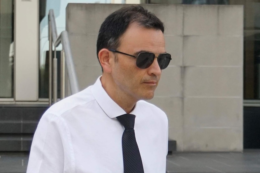 A mid-shot of a man wearing a white business shirt, black tie and sunglasses outside court.