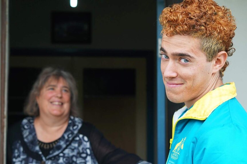 A woman with grey hair stands smiling in the background and a young man with orange dyed hair stands smiling in the foreground.