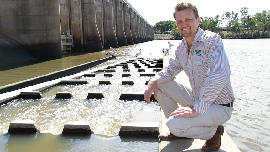 A man crouching near a fishway in a dam, with a concrete dam wall on the left.