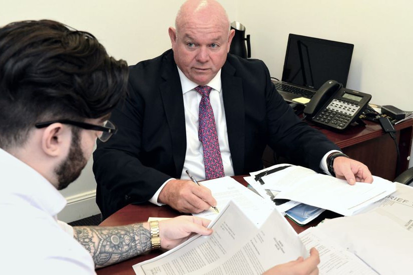 man in suit and tie sitting at table with another man as they look through documents