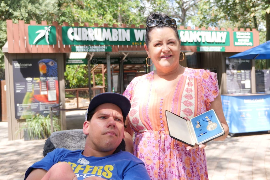 Man in wheelchari with hat and woman holding medals stand outside the entrance to the Currumbin Wildlife Sanctuary
