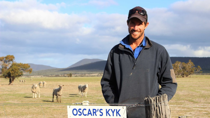 A farmer wearing a hat stands in a paddock with sheep in the background and a blue sky.