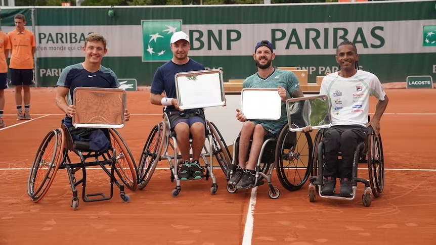 Four men in wheelchairs smile after a match at the French Open. They hold trophies in the shape of rectangular dish on the clay.