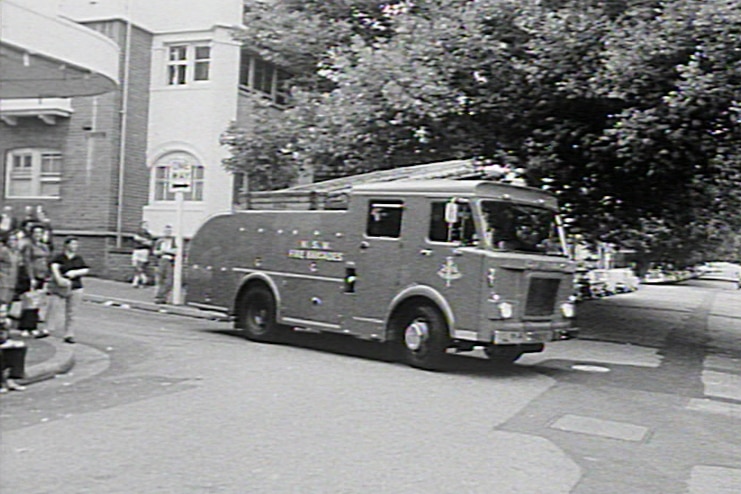 A black and white photo of a crowd of people watching a fire truck drive onto a residential street.
