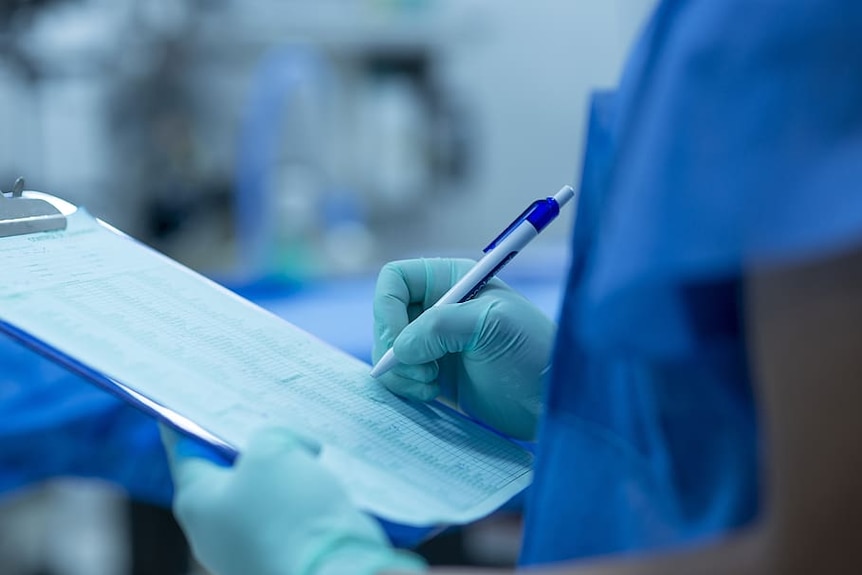 A medical worker in scrubs writes on a clipboard while wearing medical gloves.
