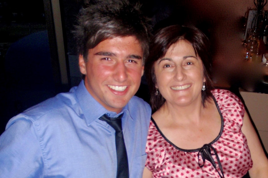 A young man in a blue shirt and black tie, smiles for a photo with his mother in a pink blouse