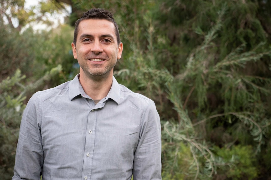 Man in grey shirt stands in front of greenery, smiling