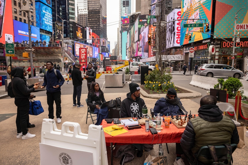 three people sit around a table with a chess board on it in Times Square, multiple plasma screens in the background. 