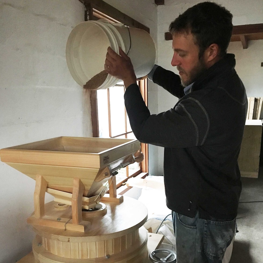 A farmer lifts a bucket of wheat into the opening of a small timber flour mill