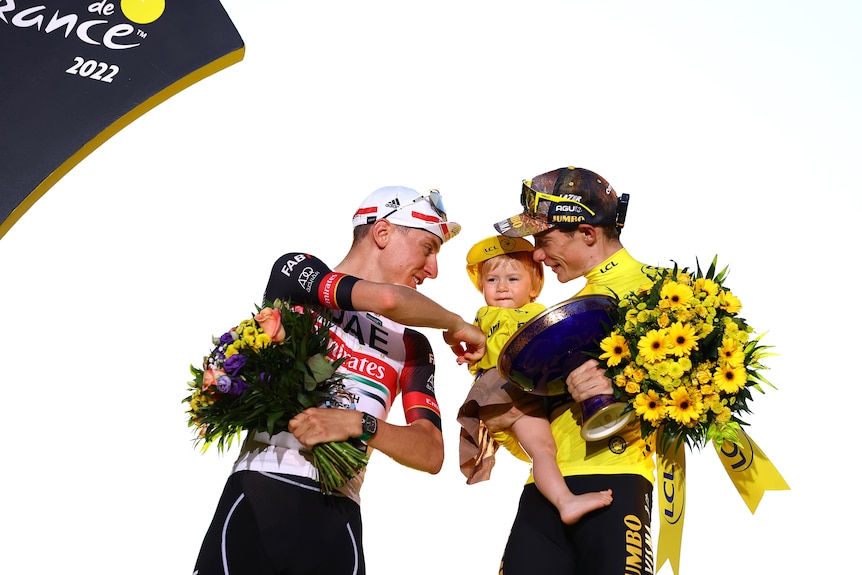 Two cyclists smile and laugh on the Tour de France podium as they look at a little girl in the arms of the yellow jersey winner.