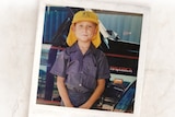 boy in school uniform with yellow hat and blue collared shirt stands in front of utility vehicle 