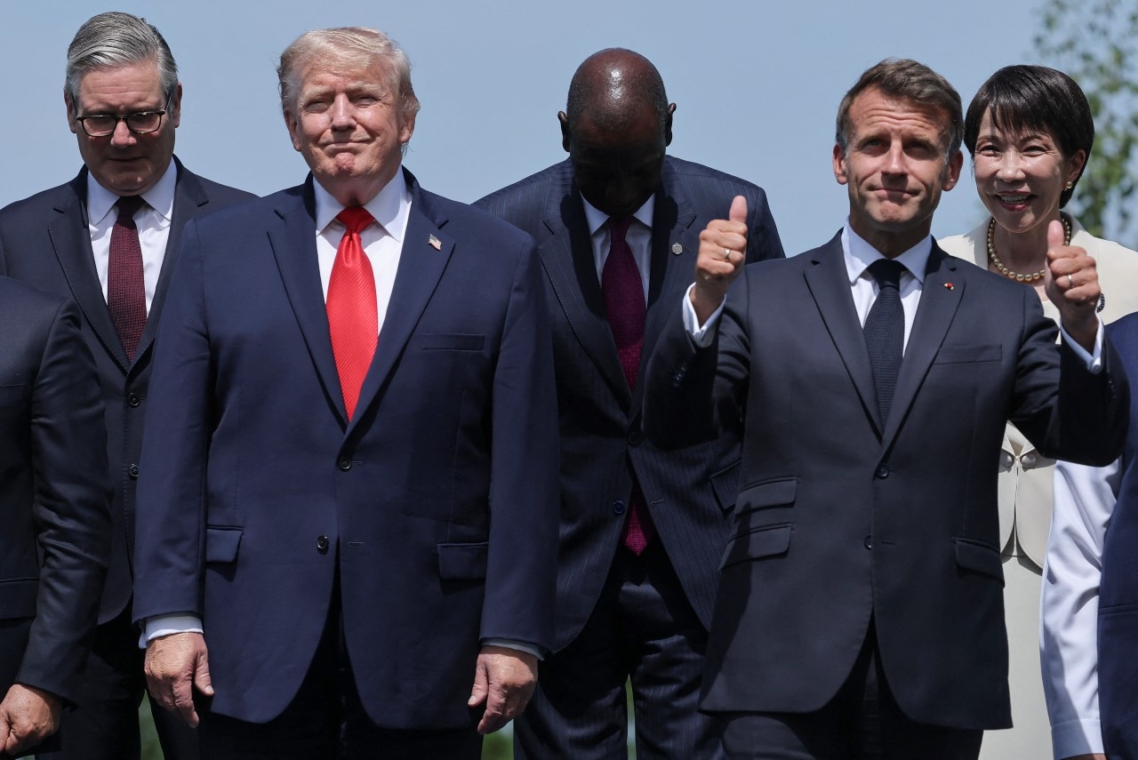 Several people on a stage including one in a suit (the French president) giving two thumbs up.