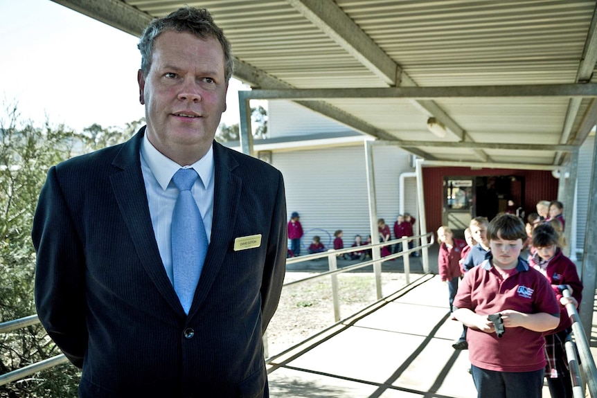 A mann in a suit with a white shirt and blue tie, with primary school students behind him