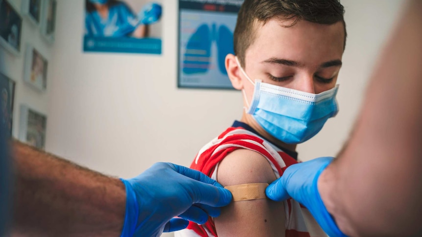 A boy wearing a blue surgical mask holds still as a nurse puts a plaster on his arm after he gets a COVID-19 jab.