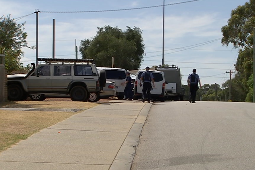 A long shot of a suburban street with white, unmarked police vans parked outside a house and officers walking around.