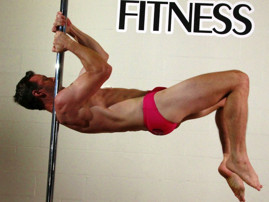 Man wears pink speedos and wraps his hips around a vertical silver pole