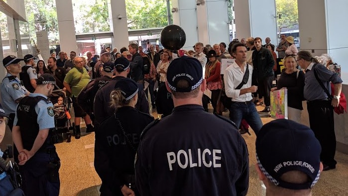 8 police officers stand facing dozens of people gathering in the foyer of a city building.