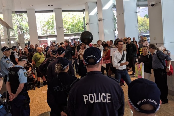 8 police officers stand facing dozens of people gathering in the foyer of a city building.