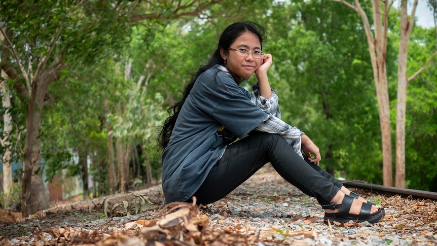 An olive-skinned young woman with long black hair + glasses sits on an old traintrack. She rests her chin on her palm, smiling