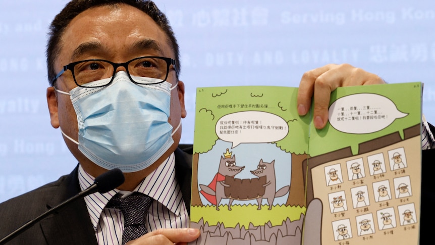 A man at a press conference holds open a children's book featuring cartoons of wolves and sheep.