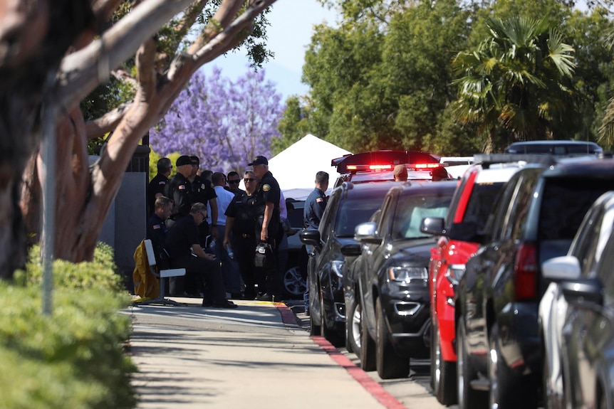 Emergency services gather on a footpath on a street lined with cars.