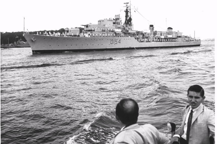 Black and white photo of navy ship on water, two men in suits men watching it from foreground. 