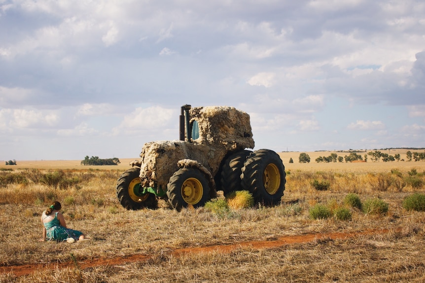 A girl sits on the ground in a dry paddock in front of a large tractor covered in wool.