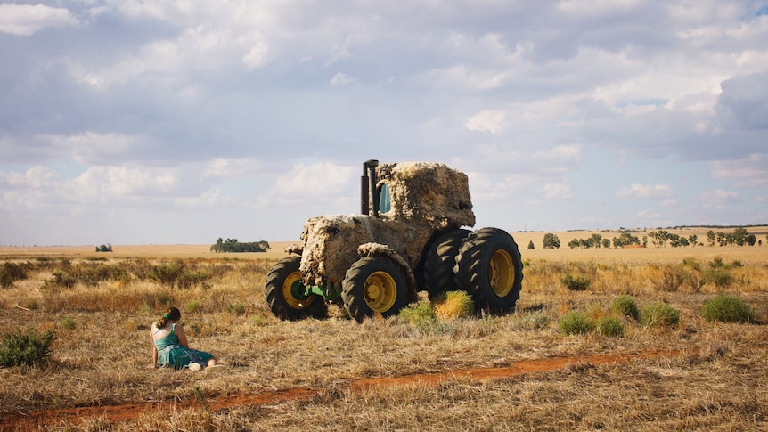 A girl sits on the ground in a dry paddock in front of a large tractor covered in wool.