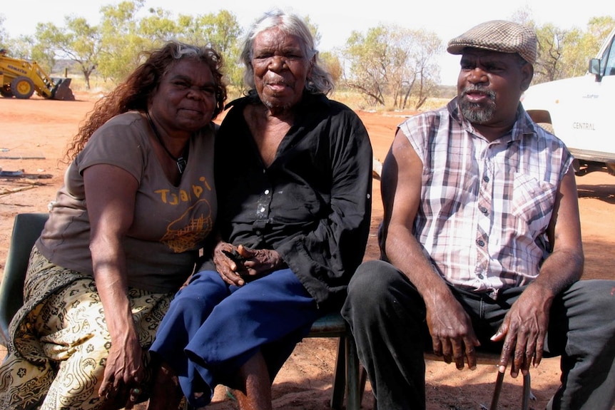 Three people sit and smile to the camera. They are outside, and there is red dirt behind them.