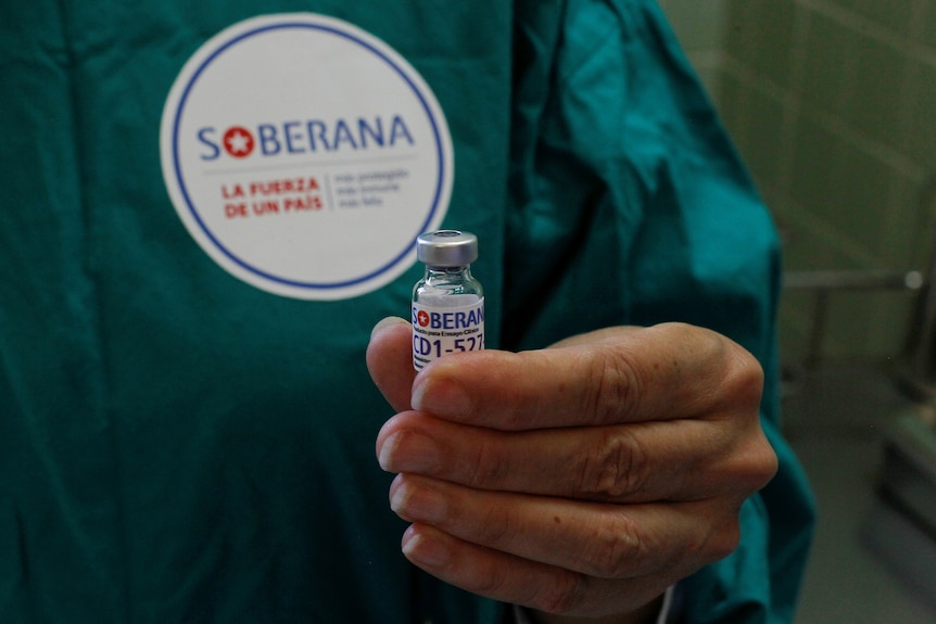 A medical worker wearing a green outfit a sticker for Soberana vaccine on it holds a vaccine vial 