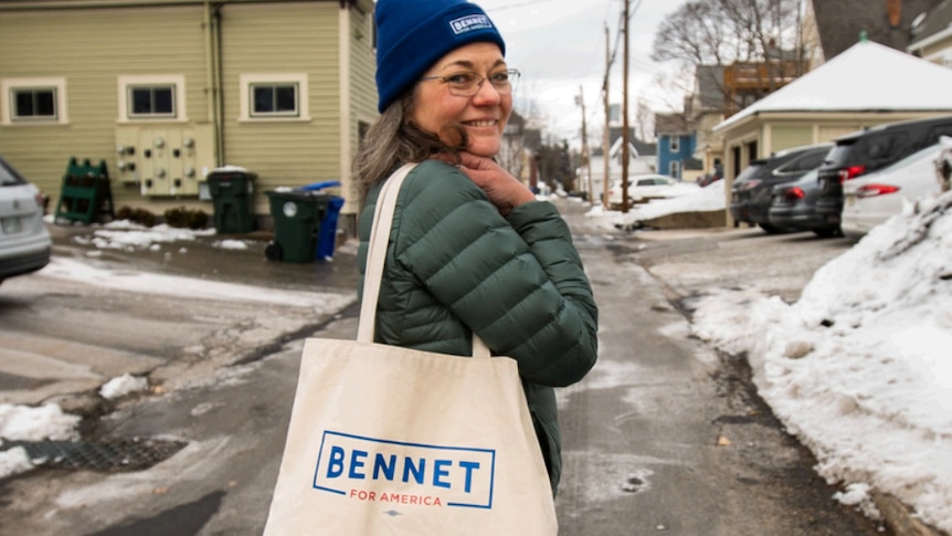 A woman stands in the road smiling as she holds a canvas bag with Bennet for America written on it