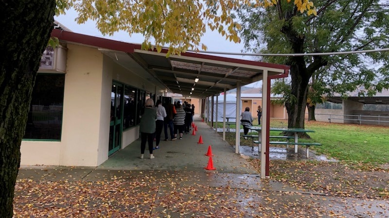 Side of building with veranda. people lined up next to orange witches hats under veranda